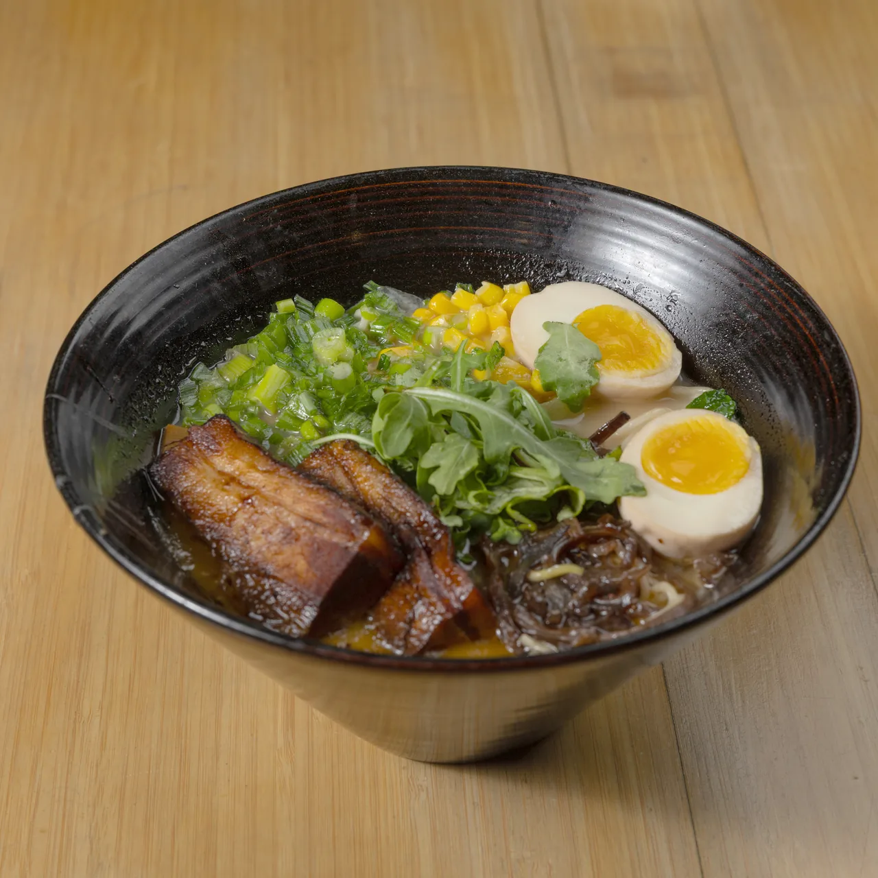 Tonkotsu ramen with braised pork belly, soft eggs, corn and arugula in a black bowl