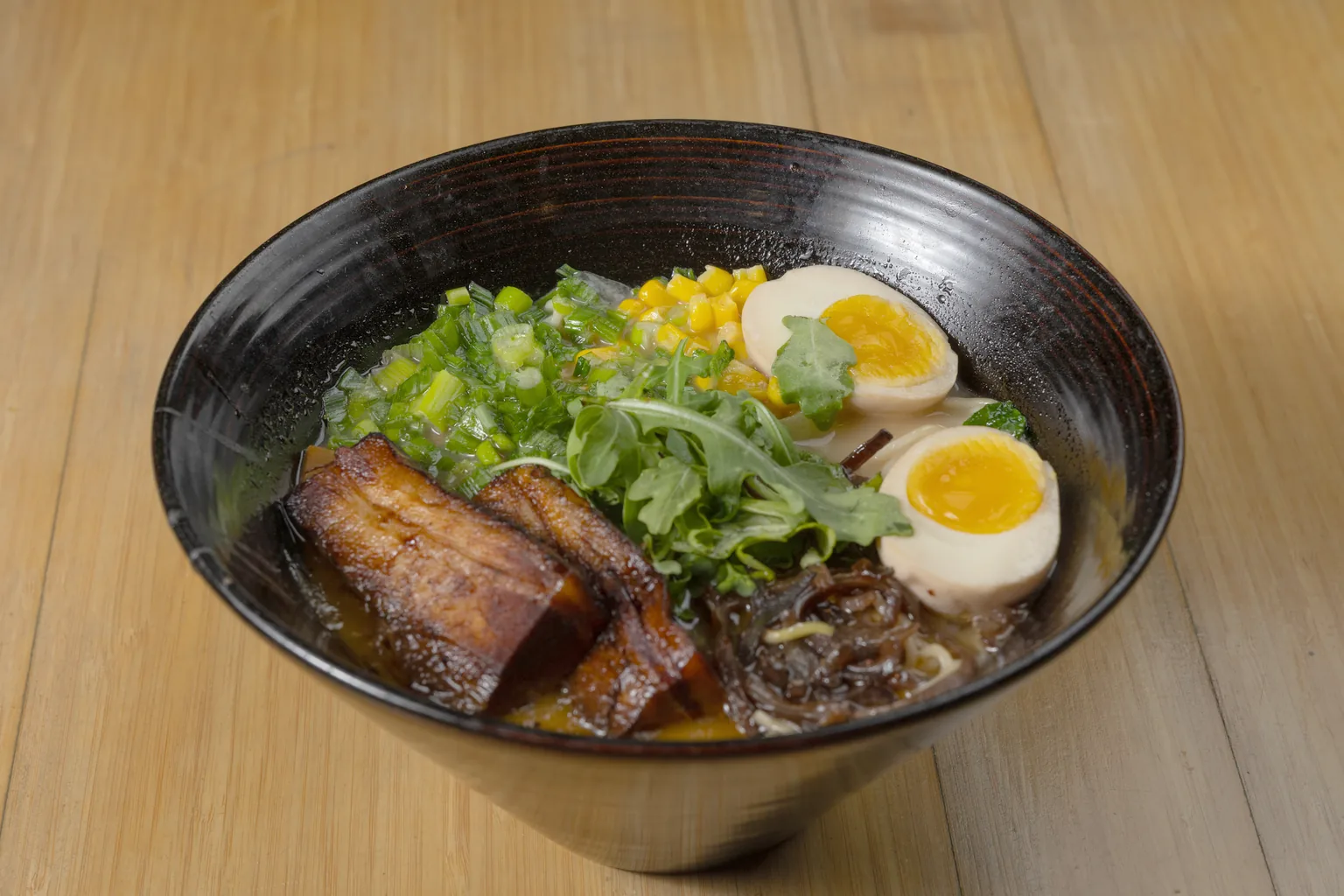 Tonkotsu ramen with braised pork belly, soft eggs, corn and arugula in a black bowl