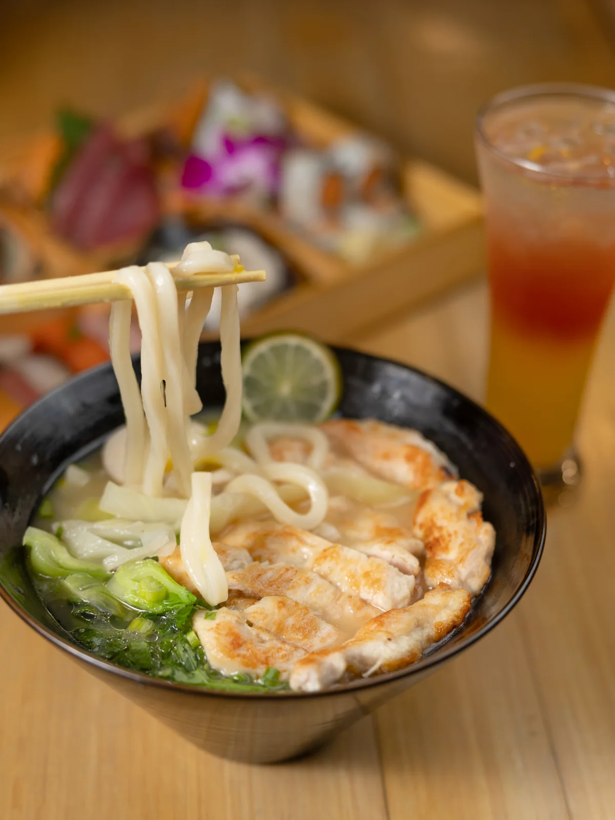 Chopsticks lifting thick noodles from a bowl of grilled chicken noodle soup